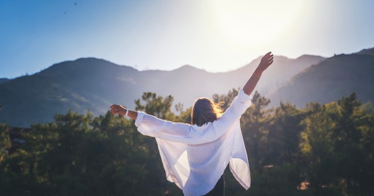 A woman in a white shirt, her hands raised, is enjoying the sun that is peeking behind the mountains.