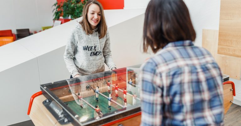 Two women are smiling and playing foosball together at a clear-top table in a bright, welcoming room.