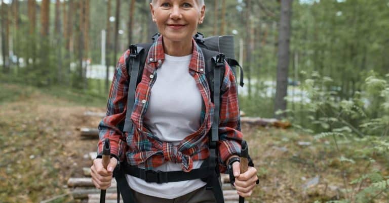 A close-up showing a middle aged woman in a flannel shirt hiking down a woodsy trail using trekking poles.