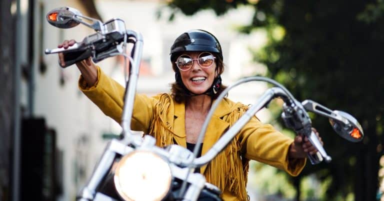 A woman wearing large sunglasses, a black helmet, and a yellow jacket smiles widely while riding a motorcycle.