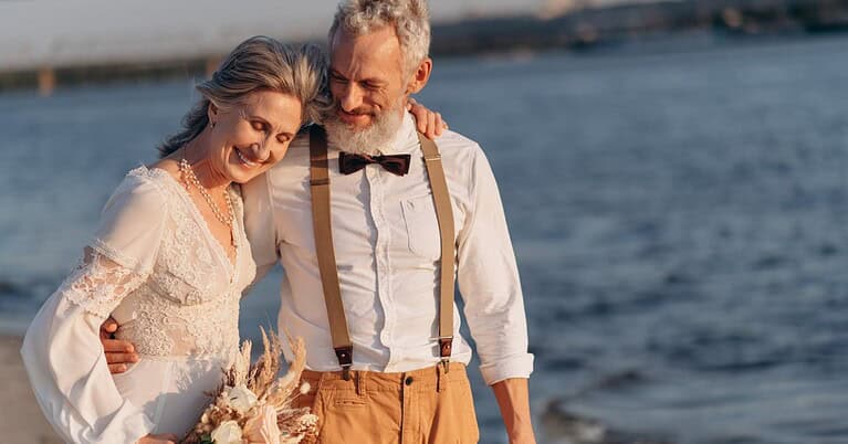 An older woman and an older man with gray hair dressed in wedding clothes embrace in front of the ocean.