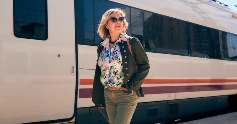 A woman wearing sunglasses and a floral shirt is carrying a suitcase while walking past a white train.