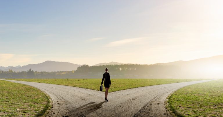 A woman in a skirt and suit blazer carrying a briefcase and standing in the middle of a forked road.