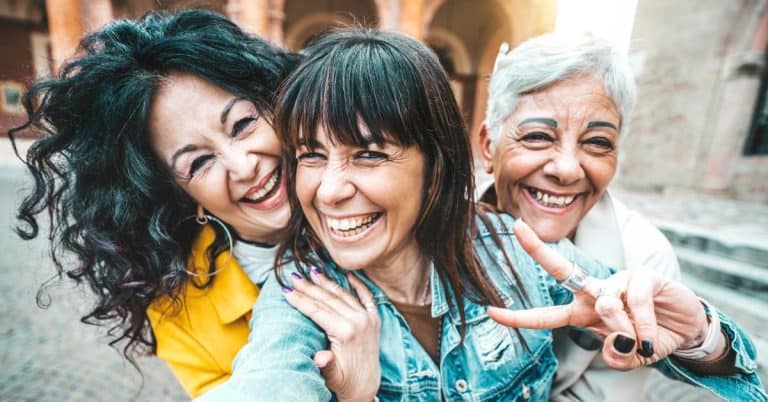 A group of middle-aged and senior women embrace and smile while taking a selfie on a city street.