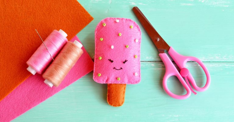 A pink popsicle craft made out of felt with an orange stick and a face sits on a table with pink scissors and thread.