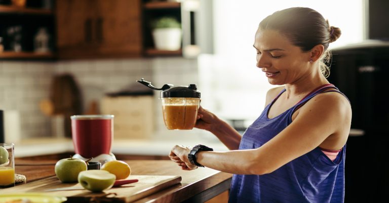 A brunette woman in a blue workout tank top holds a smoothie in the kitchen as she checks out her smartwatch.