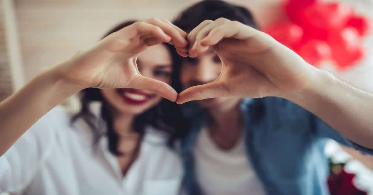 A close-up of two people creating a heart with their hands. A man and woman stand next to each other, blurred in the background.