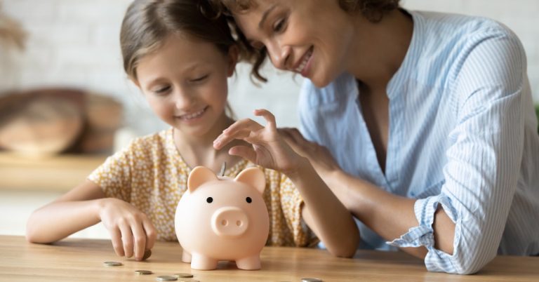 A middle-aged mom sitting at the table with her young daughter. They're putting coins in a piggy bank.