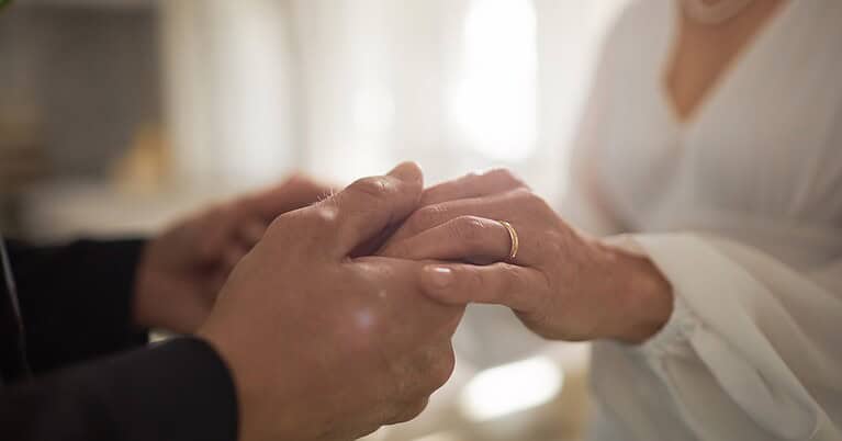 A close-up of a couple holding each other's hands. One person is wearing a flowing white dress and a gold wedding band.